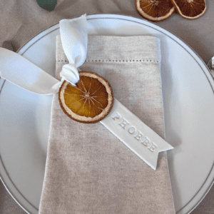 white plate, beige napkin with dried orange and white ribbon Christmas place setting name tag